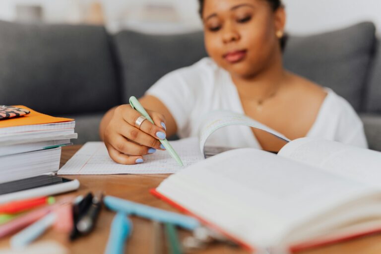 Young woman writing in a notebook with study materials around her, focused on learning at home.